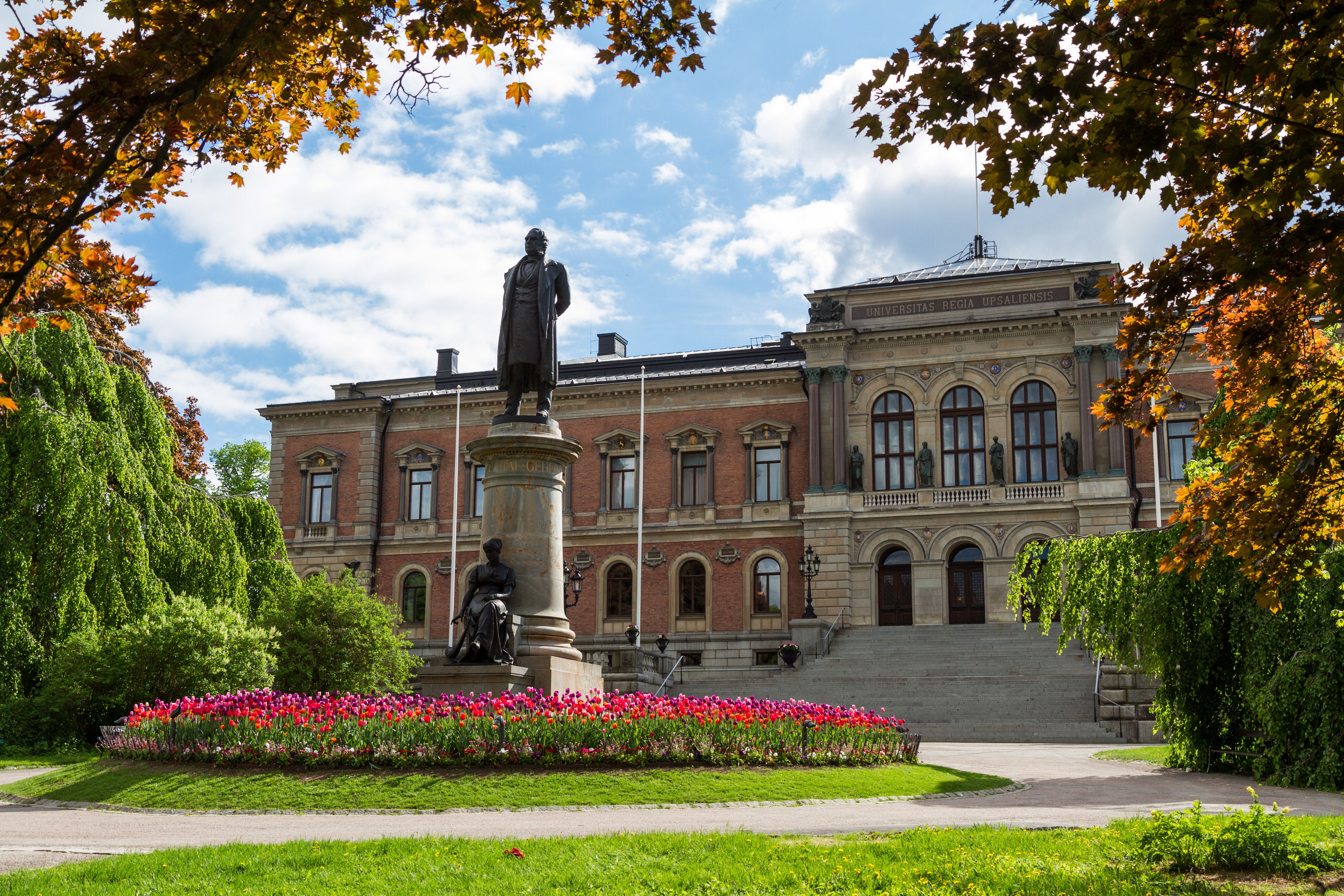 A statue, flowers and trees, in front of a 19th century building. Photo.